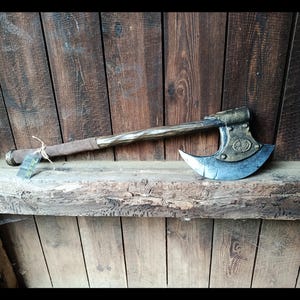 Könnte beinhalten: Eine Doppelaxt mit einem verwitterten Metallkopf mit kreisförmigem Emblem, einem gedrehten Holzstiel und einem braunen, lederummantelten Griff. Ein kleines rechteckiges Etikett ist mit einer Schnur am Stiel in der Nähe der Basis befestigt.