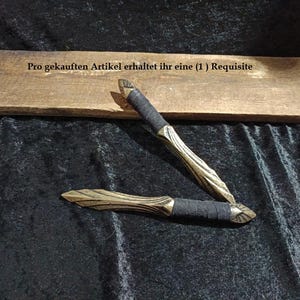 Könnte beinhalten: Zwei doppelseitige Wurfdolche mit metallisch goldfarbenen Klingen, die ein strukturiertes, blattartiges Muster aufweisen, und schwarz umwickelten Griffen. Der Text "Pro gekauften Artikel erhaltet ihr eine (1) Requisite" ist auf dem Holzbrett zu sehen.