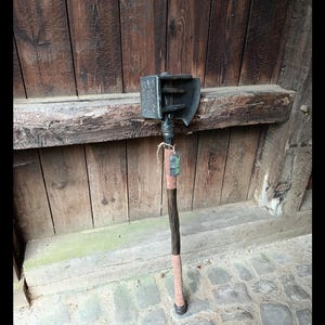 Könnte beinhalten: Ein dunkelgrauer Metall-Kriegshammer mit einem quadratischen Kopf, der ein kreisförmiges Emblem auf der Seite und eine geschwungene Klinge auf der Rückseite aufweist. Der Stiel besteht aus dunklem Holz mit hellbraunen Lederwicklungen.