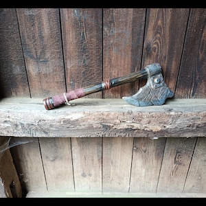 Könnte beinhalten: Eine Handaxt mit einem dunklen Holzgriff, einem kastanienbraunen Wickelgriff und metallischen Akzenten. Der Axtkopf besteht aus strukturiertem grauem Metall mit einem eingravierten Kreuzschraffurmuster und einem runden Nietendetail.