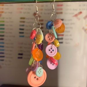 May include: A pair of dangling earrings featuring a cascade of colorful buttons in various sizes and shades, including pink, orange, yellow, blue, and purple. The buttons are attached to silver-tone metal hooks and rings.