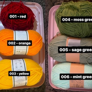May include: A selection of colorful yarn skeins in a pink tray. The colors include red, orange, yellow, moss green, sage green, and mint green. Each skein is labeled with a number and color name, ideal for knitting or crochet projects.