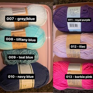 May include: A selection of colorful yarn skeins in a pink tray. Colors include grey/blue, tiffany blue, teal blue, navy blue, royal purple, lilac, and barbie pink. Each skein has a label with a number and color name.