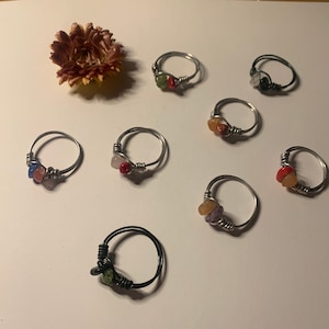 May include: A collection of handmade wire-wrapped rings with colorful gemstone accents. The rings feature silver and black wire, with stones in various colors, including red, green, and orange. A dried flower is also present.