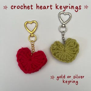 May include: Two crochet heart keyrings, one red and one olive green. The red heart has a gold-colored keyring, and the green heart has a silver-colored keyring. The text "crochet heart keyrings" and "gold or silver keyring" are visible.