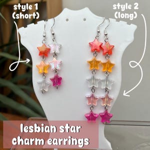 Puede incluir: Dos pares de pendientes con colgantes de estrella de diferentes longitudes. Los pendientes presentan un degradado de cuentas en forma de estrella de colores, que van del rojo al rosa, con ganchos plateados. El texto "lesbian star charm earrings" está en la parte inferior.