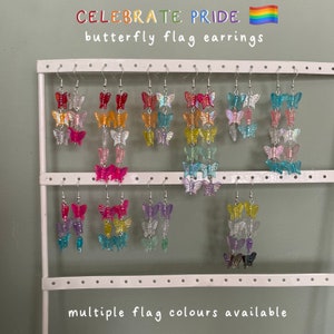 Może przedstawiać: Prezentacja kolczyków w kształcie motyli w kolorach tęczy, w tym czerwonym, pomarańczowym, żółtym, zielonym, niebieskim i fioletowym. Kolczyki wiszą na białym stojaku wystawowym. U góry napis "CELEBRATE PRIDE" i "butterfly flag earrings". Na dole napis "multiple flag colours available".