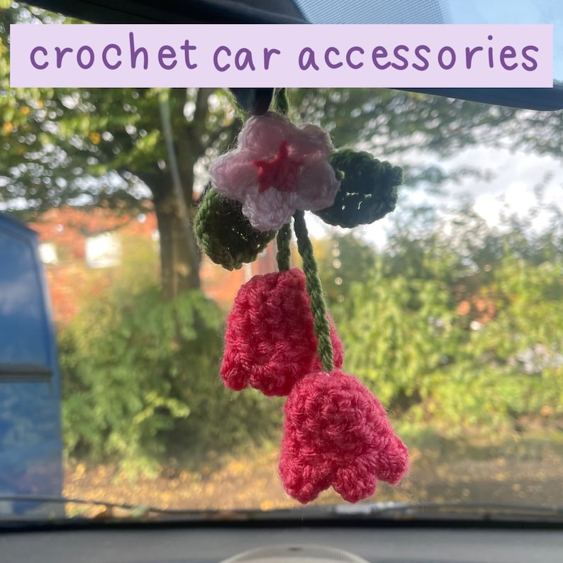 May include: Crochet car accessories. A handmade car charm featuring a pink and white flower with green leaves and two pink tulip-shaped pendants. The text "crochet car accessories" is displayed in a light purple banner.