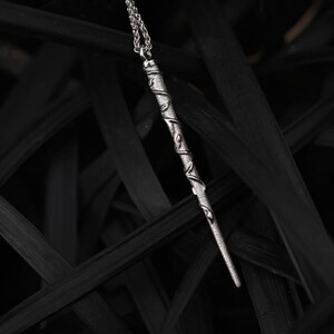 Puede incluir: Un collar de plata con un colgante en forma de varita m&aacute;gica, adornado con enredaderas y hojas. El collar est&aacute; fotografiado sobre un fondo negro.