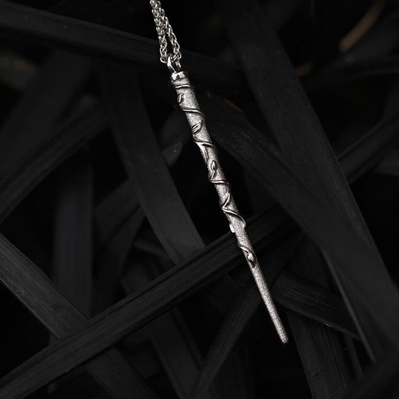 Puede incluir: Un collar de plata con un colgante en forma de varita m&aacute;gica, adornado con enredaderas y hojas. El collar est&aacute; fotografiado sobre un fondo negro.