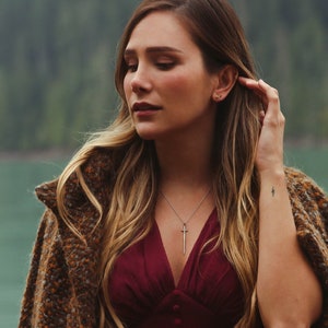 Puede incluir: Una mujer con el pelo largo y casta&ntilde;o lleva un vestido burdeos con escote pronunciado y un c&aacute;rdigan marr&oacute;n. Lleva un collar plateado con un colgante de espada. Tiene un peque&ntilde;o tatuaje de rayo en la mu&ntilde;eca.