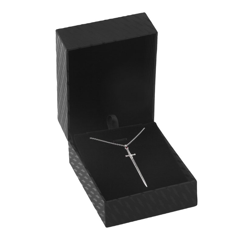 Puede incluir: Un collar con un colgante de espada de plata con una piedra roja en la empu&ntilde;adura, presentado en una caja de joyer&iacute;a de terciopelo negro.