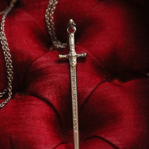Puede incluir: Un colgante de espada de plata con una joya roja en la empu&ntilde;adura. La espada est&aacute; inscrita con las palabras "Godric Gryffindor". El colgante est&aacute; en una cadena de plata y descansa sobre una superficie de terciopelo rojo.