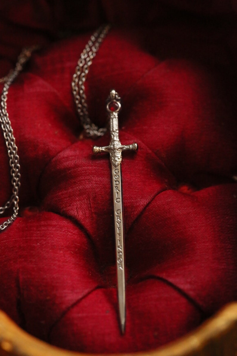 Puede incluir: Un colgante de espada de plata con una joya roja en la empu&ntilde;adura. La espada est&aacute; inscrita con las palabras "Godric Gryffindor". El colgante est&aacute; en una cadena de plata y descansa sobre una superficie de terciopelo rojo.