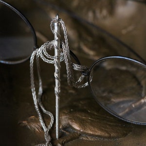 Puede incluir: Un collar de cadena de plata con un colgante de plata retorcido que cuelga de &eacute;l. El collar est&aacute; drapeado sobre un par de gafas.