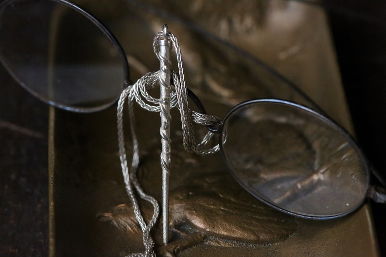 Puede incluir: Un collar de cadena de plata con un colgante de plata retorcido que cuelga de &eacute;l. El collar est&aacute; drapeado sobre un par de gafas.