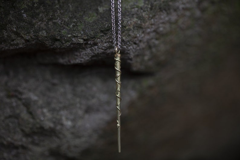 Puede incluir: Un collar de cadena plateada con un colgante de varita dorada. La varita est&aacute; detallada con un dise&ntilde;o en espiral.