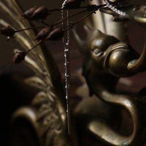 Puede incluir: Un collar de cadena de plata con un largo y delgado colgante cuelga de un candelero de lat&oacute;n. El candelero tiene forma de cabeza y cuello de p&aacute;jaro, con un dise&ntilde;o detallado.
