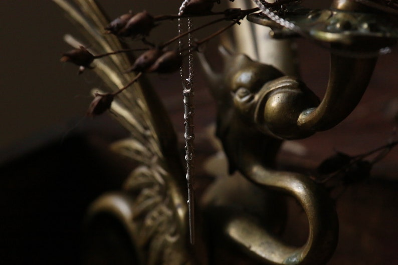 Puede incluir: Un collar de cadena de plata con un largo y delgado colgante cuelga de un candelero de lat&oacute;n. El candelero tiene forma de cabeza y cuello de p&aacute;jaro, con un dise&ntilde;o detallado.