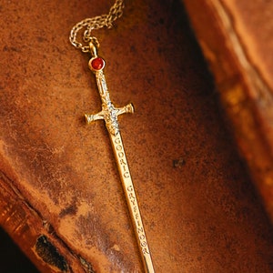 Puede incluir: Un collar de espada de oro con una piedra preciosa roja en la parte superior. La espada est&aacute; grabada con las palabras "Godric Gryffindor".