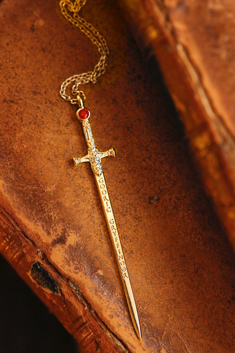 Puede incluir: Un collar de espada de oro con una piedra preciosa roja en la parte superior. La espada est&aacute; grabada con las palabras "Godric Gryffindor".