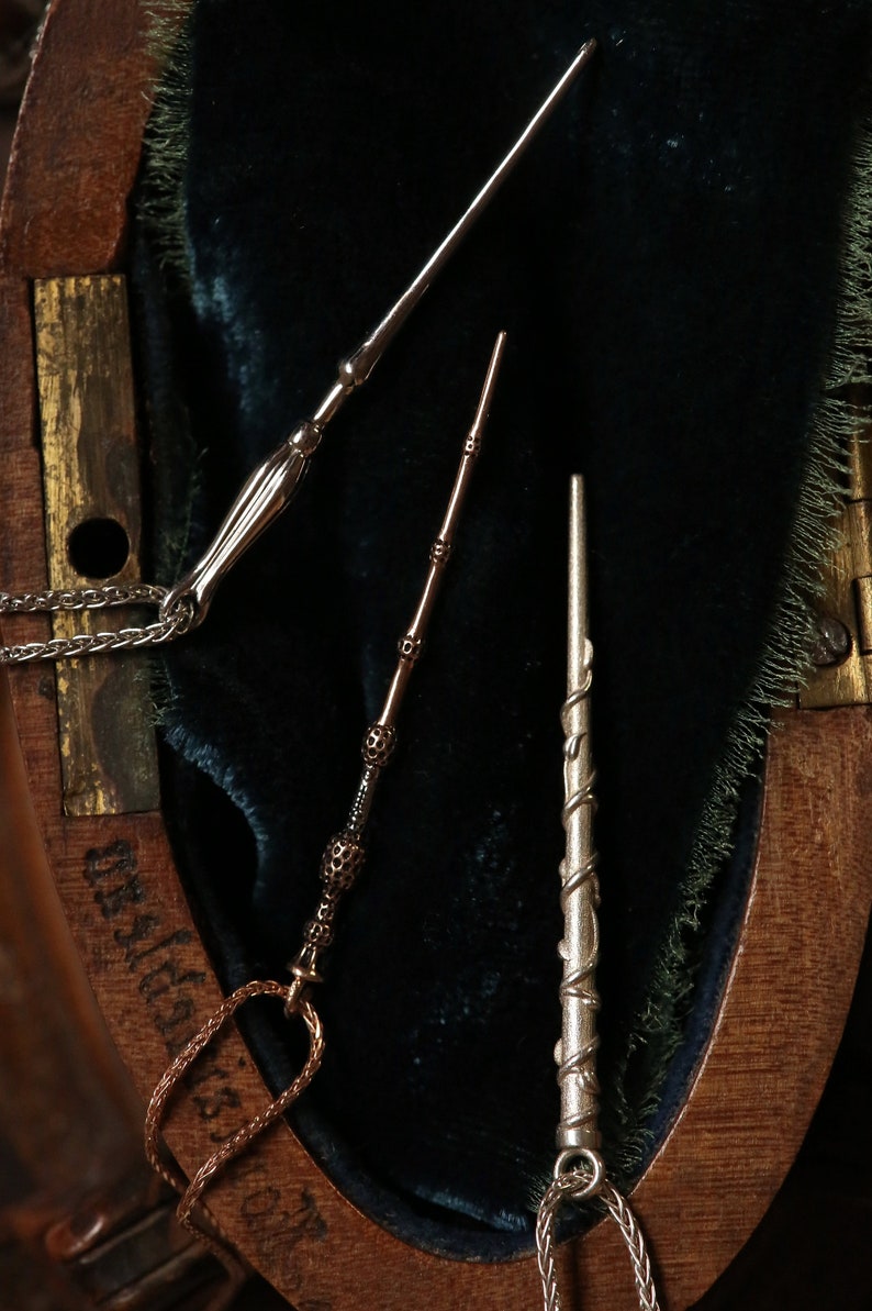 Puede incluir: Tres collares con varitas de plata de diferentes dise&ntilde;os. Las varitas est&aacute;n sobre un fondo de terciopelo azul oscuro dentro de una caja de madera.