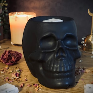May include: A black ceramic skull candle holder with a white wax melt inside. The skull has a detailed design and is sitting on a wooden surface with dried rose petals.