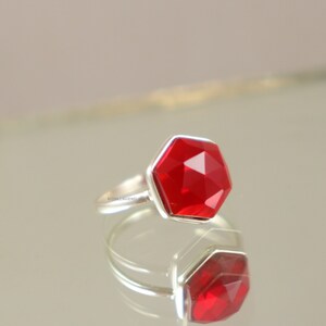 Può includere: Un anello d'argento con una pietra preziosa esagonale rossa. L'anello è su uno sfondo bianco.