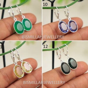 May include: Four pairs of silver dangle earrings with oval-shaped gemstones. The earrings are shown on a hand against a green background. The gemstones are green, purple, yellow, and black.