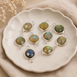 May include: A collection of round labradorite gemstone pendants with silver-colored metal settings. The gemstones display a range of colors, including green, blue, and yellow. The pendants are arranged on a white, scalloped-edge plate.