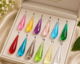 May include: A set of twelve faceted, teardrop-shaped crystals arranged in a beige display box labeled BISMILLAH JEWELLERY. Each crystal is numbered one through twelve and features a distinct color, including pink, yellow, purple, light pink, blue, green, red, teal, brown, lime, pale yellow, and clear.