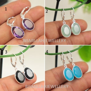 May include: Four pairs of silver dangle earrings with different colored gemstone drops. The earrings are shown on a hand against a green background. The gemstones are amethyst, green, black, and blue.