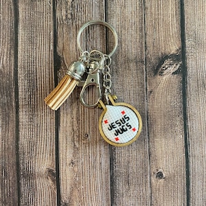 May include: A silver key chain with a brown tassel and a round wooden charm with the text "JESUS JUGS" embroidered in black thread.