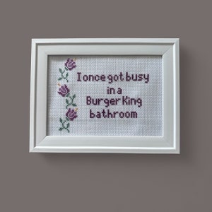 May include: White framed cross-stitch with purple flowers and the text "I once got busy in a Burger King bathroom."