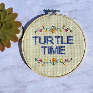 May include: A cross-stitch embroidery hoop with the words "TURTLE TIME" in blue. The design is on a cream-colored fabric, with floral accents in pink, orange, and green. The wooden hoop is visible, and a succulent is in the background.