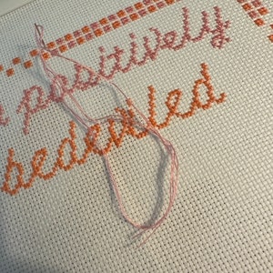 May include: Close-up of a cross-stitch project with the phrase "positively bewildered" stitched in pink and orange thread on white Aida cloth.