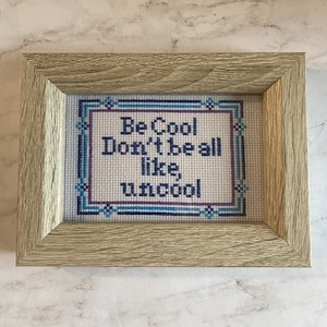 May include: A framed cross-stitch with a blue and white border and the text "Be Cool Don't be all like, uncool" in black.