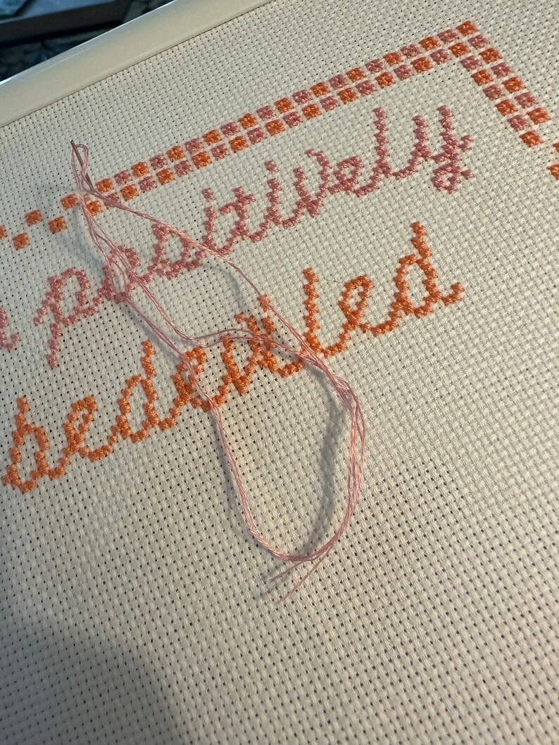 May include: Close-up of a cross-stitch project with the phrase "positively bewildered" stitched in pink and orange thread on white Aida cloth.