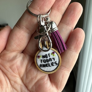 Op de afbeelding: Een sleutelhanger met een paarse kwast, een zilveren sluiting en een kleine houten cirkel met de geborduurde tekst "Not Today Ankles".