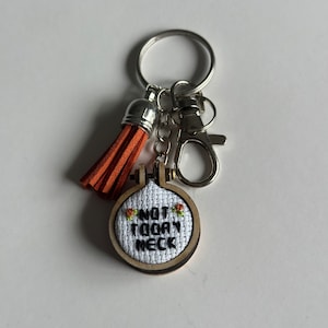 Op de afbeelding: Een kleine houten sleutelhanger met een witte stoffen cirkel waarop "NOT TODAY" in zwarte borduurwerk staat. De cirkel is omgeven door een bruin houten frame. De sleutelhanger heeft een zilveren metalen ring en een bruine kwast.