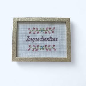 May include: A framed cross-stitch piece with the word "Ingredientses" in purple thread. The text is centered on a light blue background, framed by a gold glittery border. Floral designs in pink, green, and blue accent the top and bottom.