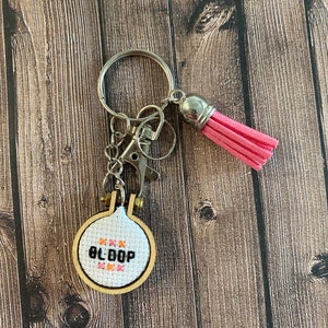 May include: A white and pink cross-stitch keychain with the word "BLOOP" stitched in black thread. The keychain is attached to a silver key ring with a pink tassel.