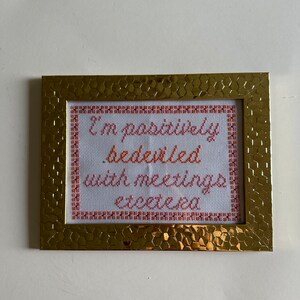May include: A gold-colored frame with a white background and cross-stitched text that reads "I'm positively bedecked with meetings etcetera".