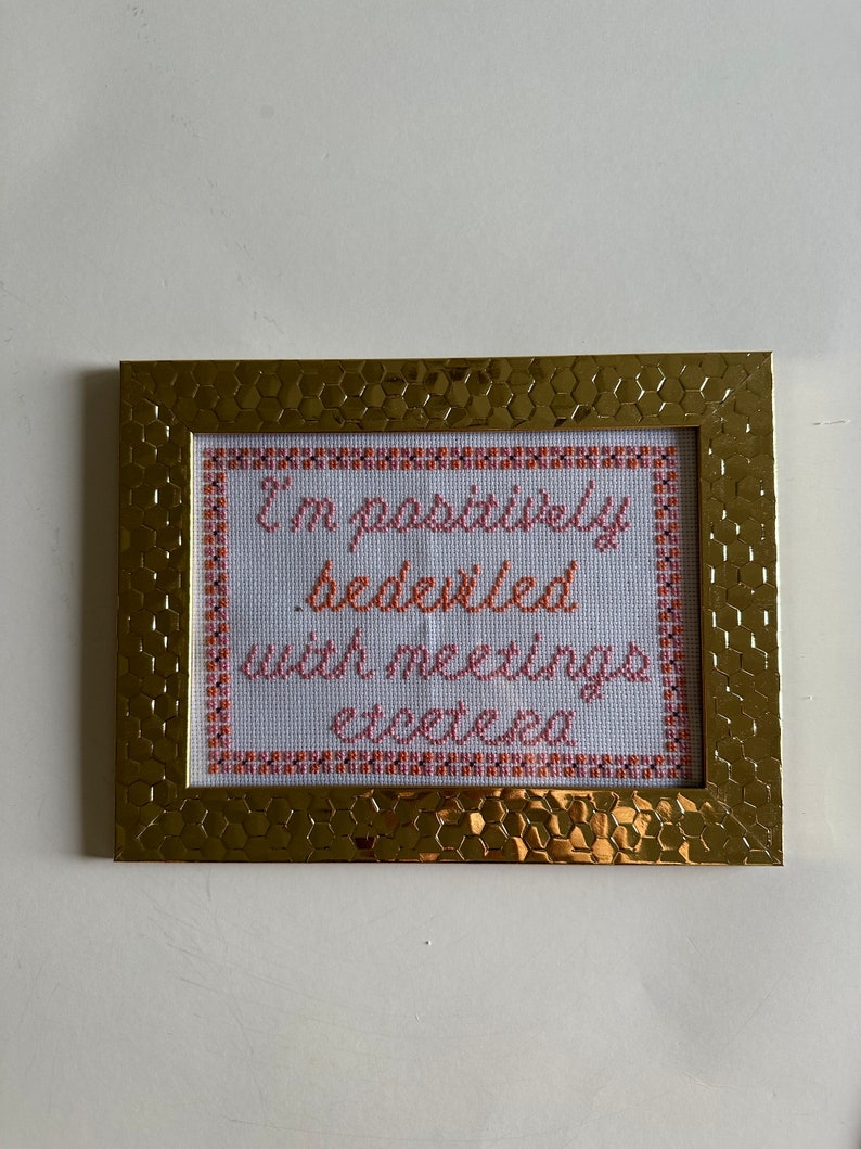 May include: A gold-colored frame with a white background and cross-stitched text that reads "I'm positively bedecked with meetings etcetera".
