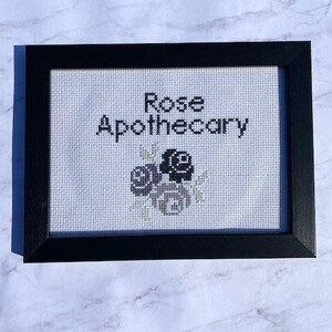 Può includere: Un ricamo a punto croce incorniciato con rose ricamate in nero e il testo "Rose Apothecary".