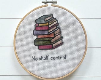 May include: Cross-stitch embroidery in a wooden hoop features a stack of colorful books. The text "No shelf control" is stitched below the books. The embroidery is on a white fabric background.