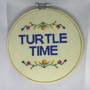 May include: A white embroidery hoop with a beige fabric background. The fabric has a blue embroidered phrase "TURTLE TIME" with a green vine and orange and yellow flowers above and below the phrase.