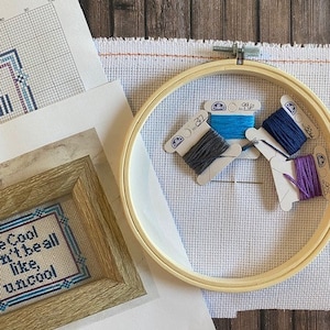 Könnte beinhalten: Nahaufnahme eines Kreuzstichprojekts mit einem hölzernen Stickrahmen und vier Spulen Stickgarn in Blau, Lila, Grau und Dunkelblau. Das Kreuzstichprojekt sagt "Be Cool Don't Be All Like Uncool".