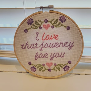 May include: A white embroidery hoop with a purple and green floral design around the edge. The text "I love that journey for you" is embroidered in the center in pink and purple thread.