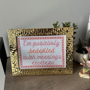 May include: A gold-colored frame with a white background and orange and pink text that reads "I'm positively bedeviled with meetings, etcetera."